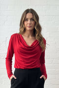 A woman stands in a bright room with a white brick wall, wearing a bright red merino wool long-sleeve cowl-neck top and black wide-leg pants.