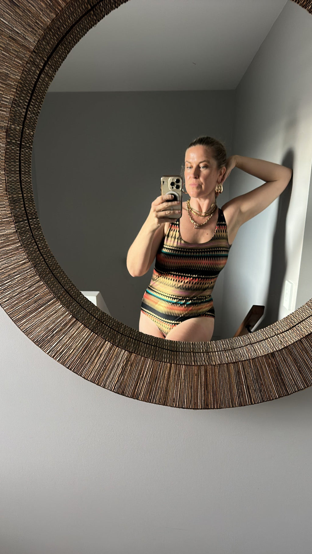 A woman takes a mirror selfie in a bathroom wearing a colorful striped one-piece swimsuit. The product shown is the swimsuit, which features a multicolor pattern of orange, yellow, green, and blue stripes on a dark background.