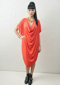A woman modeling a bright coral-red draped short-sleeve dress with a deep cowl-neckline, styled with a chunky gold chain necklace and black ankle boots against a white brick wall background.