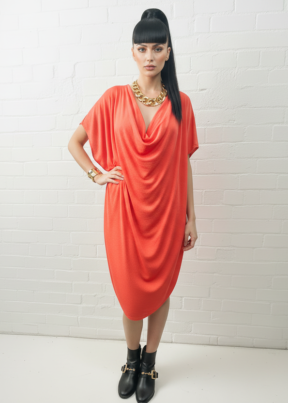 A woman modeling a bright coral-red draped short-sleeve dress with a deep cowl-neckline, styled with a chunky gold chain necklace and black ankle boots against a white brick wall background.